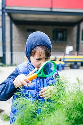 Ein Junge beobachtet mit ener großen grünen Lupe Pflanzen an einem Hochbeet