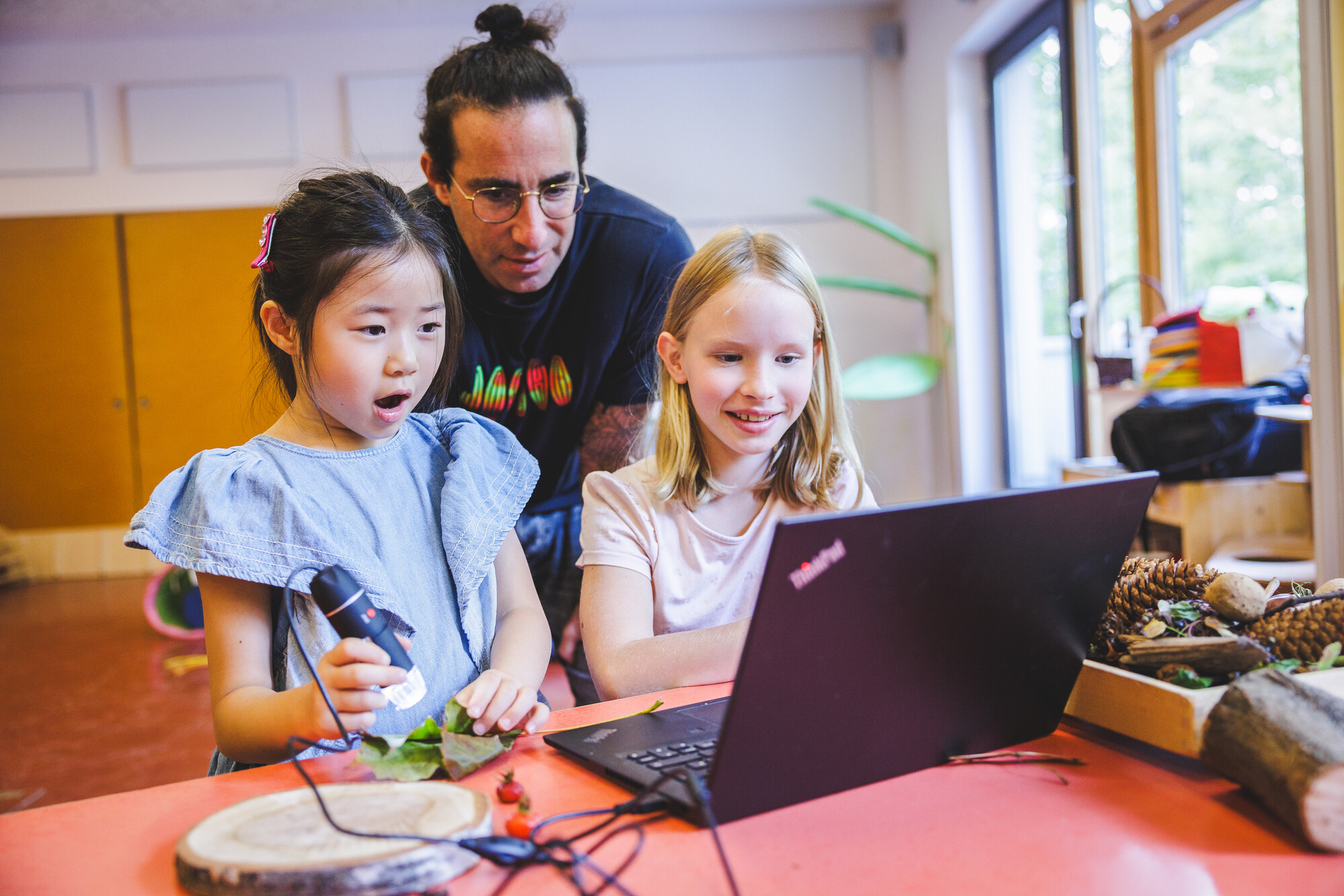 Ein Mädchen untersucht mit einem digitalen Mikroskop ein Blatt und sieht das Ergebnis zusammen mit ihrem Lehrer und einem weiteren Mädchen auf dem Laptop.