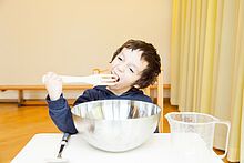 Ein Junge sitzt an einem Tisch mit übergroße Geschirr. Er versucht die große Gabel in den Mund zu nehmen. 
