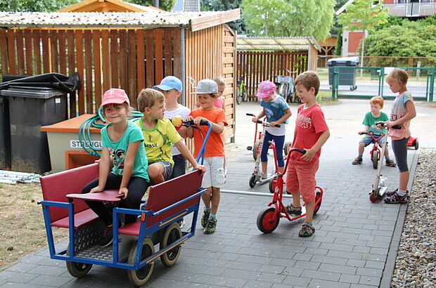 Eine Gruppe Kitakinder auf dem Weg mit einem Rollwagen und auf Rollern