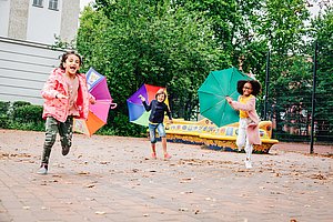 Drei Kinder rennen draußen mit bunten Regenschirmen