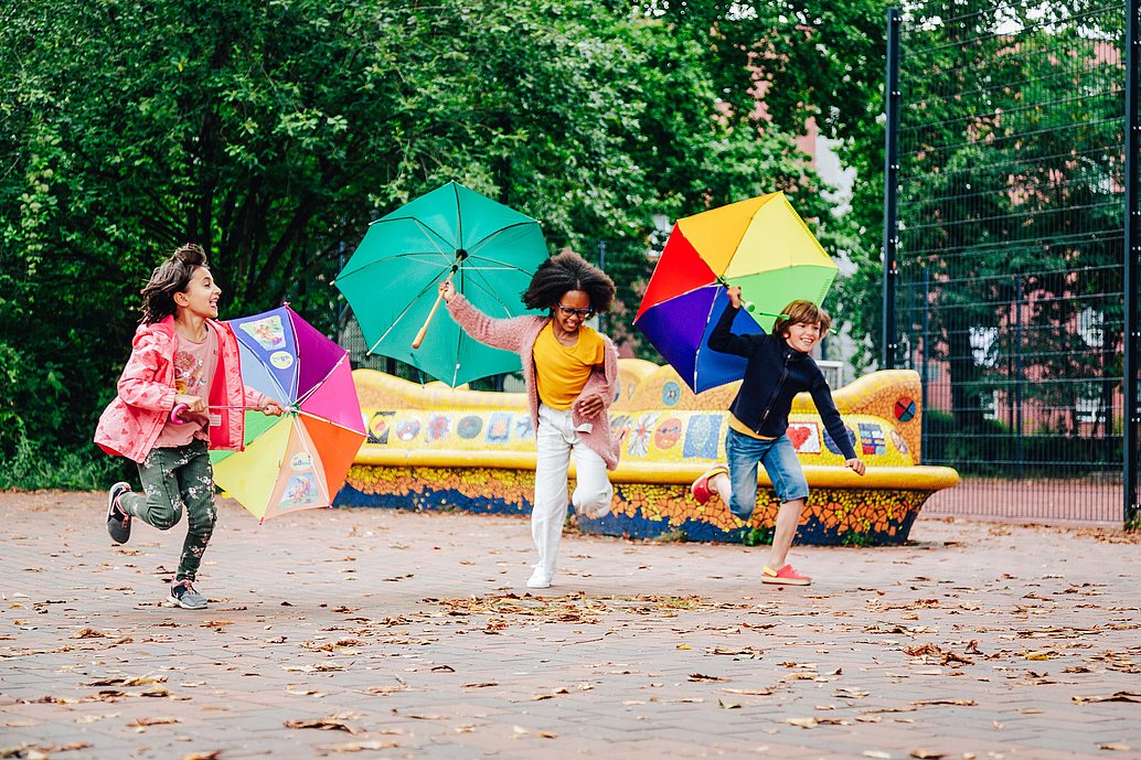 Kinder rennen mit offenen Regenschirmen fröhlich über einen Spielplatz