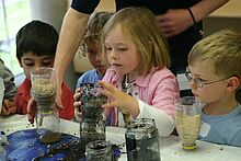 Mehrere Kinder sitzen am Tisch vor mit Schmutz bedeckten Gläsern und zerschnittenen Wasserflaschen