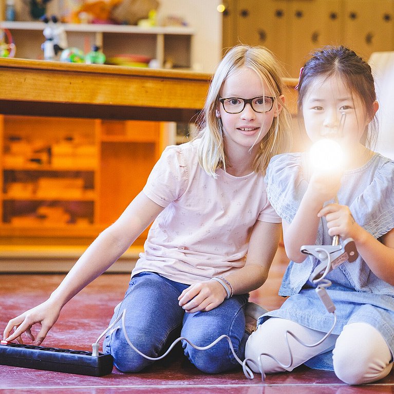 Zwei Mädchen knien auf dem Boden und leuten mit einer Lampe Richtung Kamera.