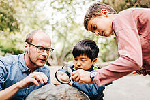 Zwei Kinder und ein Mann untersuchen mit einer Lupe einen großen Stein