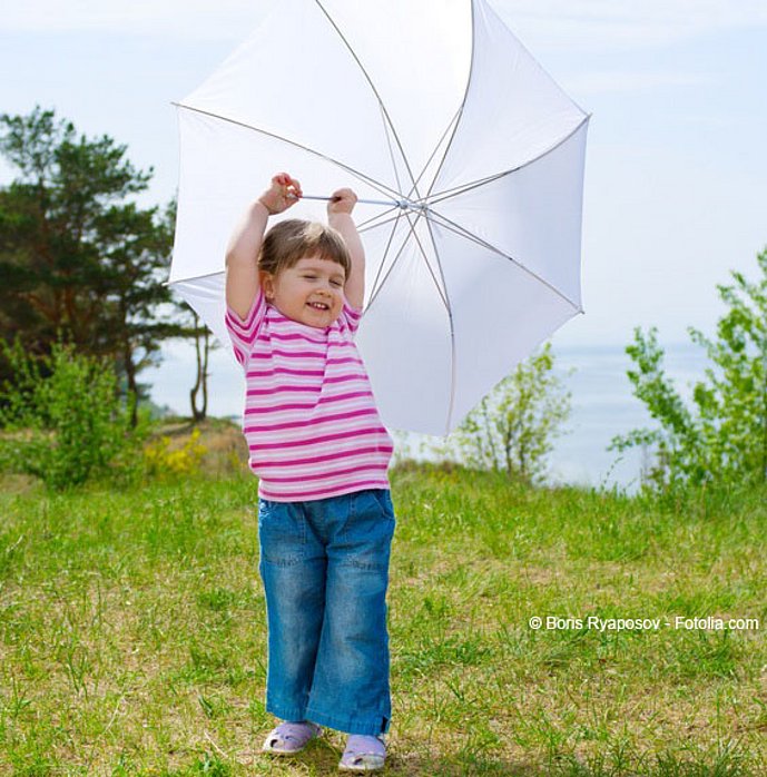 Junges Mädchen steht mit offenem weißem Regenschirm in einer Wiese.