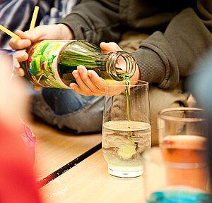 Jemand gießt eine grüne Flüssigkeit aus einer Glasflasche in ein mit einer durchsichigen Flüssigkeit gefülltes Glas