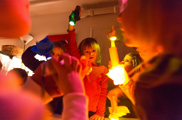 Mehrere Kinder mit bunten Taschenlampen stehen in einem dunklen Raum.