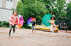 Drei Kinder rennen mit bunten aufgespannten Regenschirmen über einen Hof auf die Kamera zu. Auf dem Boden liegt Laub.
